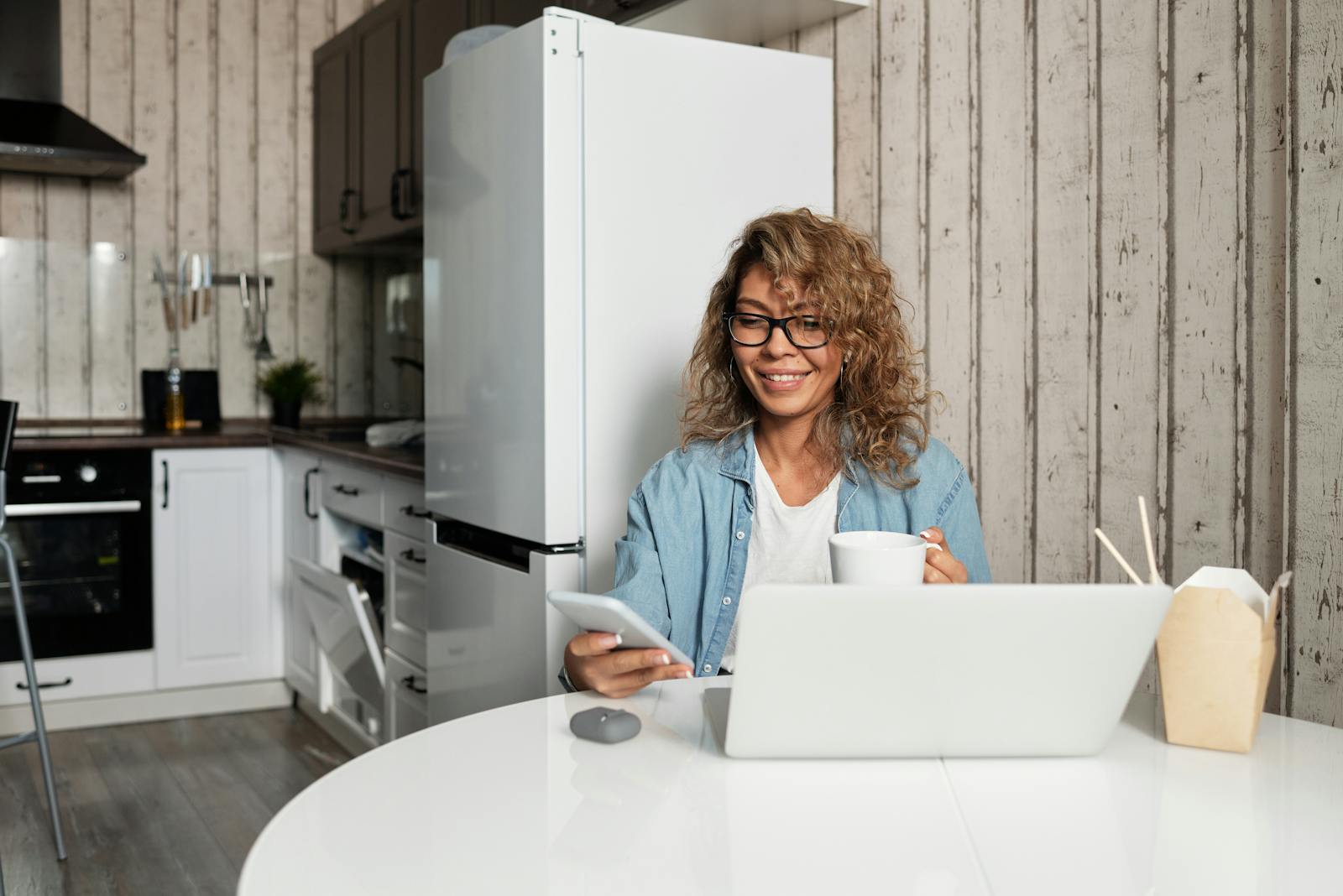 A woman using a phone and laptop while enjoying coffee at home in a rustic kitchen.
